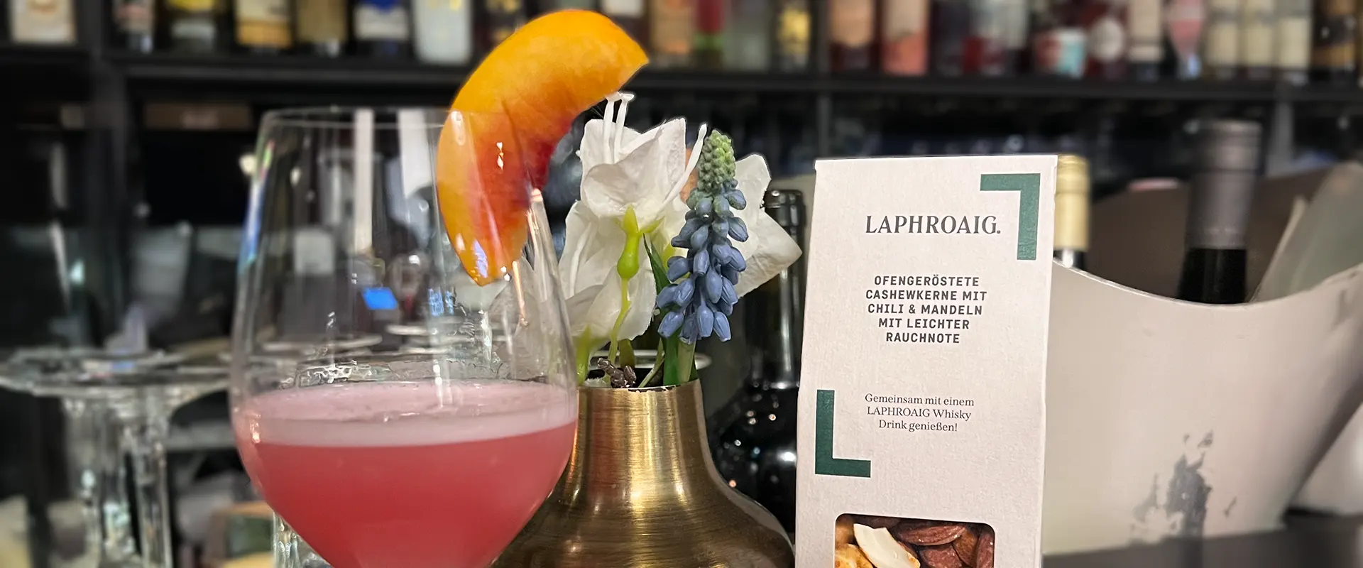Platzhalter_1920x800 Ein pinkfarbener Cocktail mit einer Orangenscheibe, neben einem Glas mit Nüssen und einer kleinen Blumenvase in der Cole & Porter Bar.