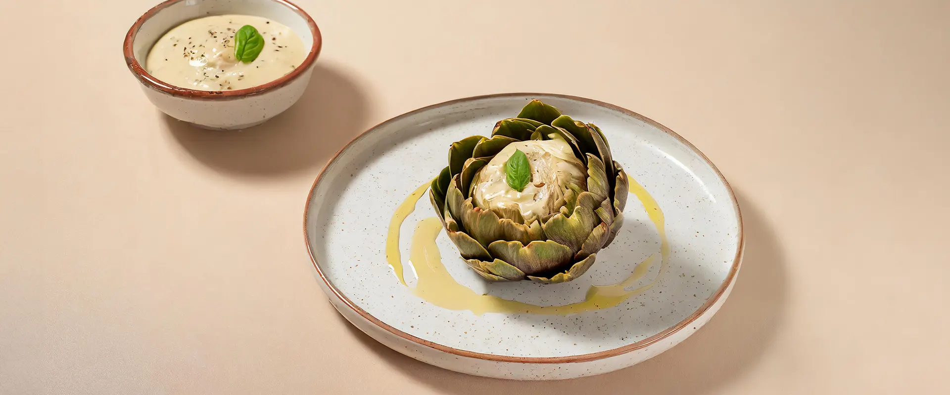 Platzhalter_1920x800 Eine ganze, gegarte Artischocke mit herzhaften Blättern, serviert auf einem Teller mit einer kleinen Schale Dip, garniert mit einem frischen Basilikumblatt.