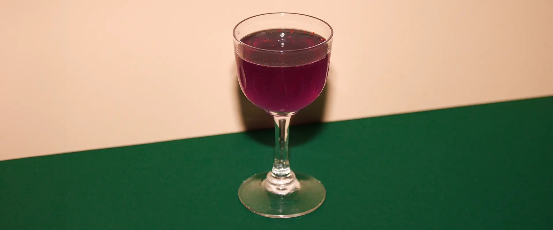 Platzhalter_1920x800 Ein Glas mit einem lila Cherry Lady Getränk steht auf einem grünen Untergrund vor einem beigefarbenen Hintergrund.