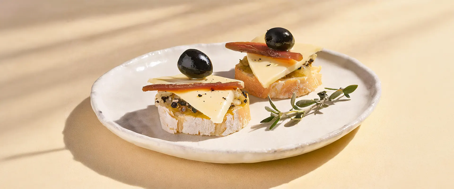 Platzhalter_1920x800 Zwei herzhafte Canapés auf einem weißen Teller. Jede Scheibe besteht aus einem Stück Brot, belegt mit Käse, einer schwarzen Olive und einer Saredelle.