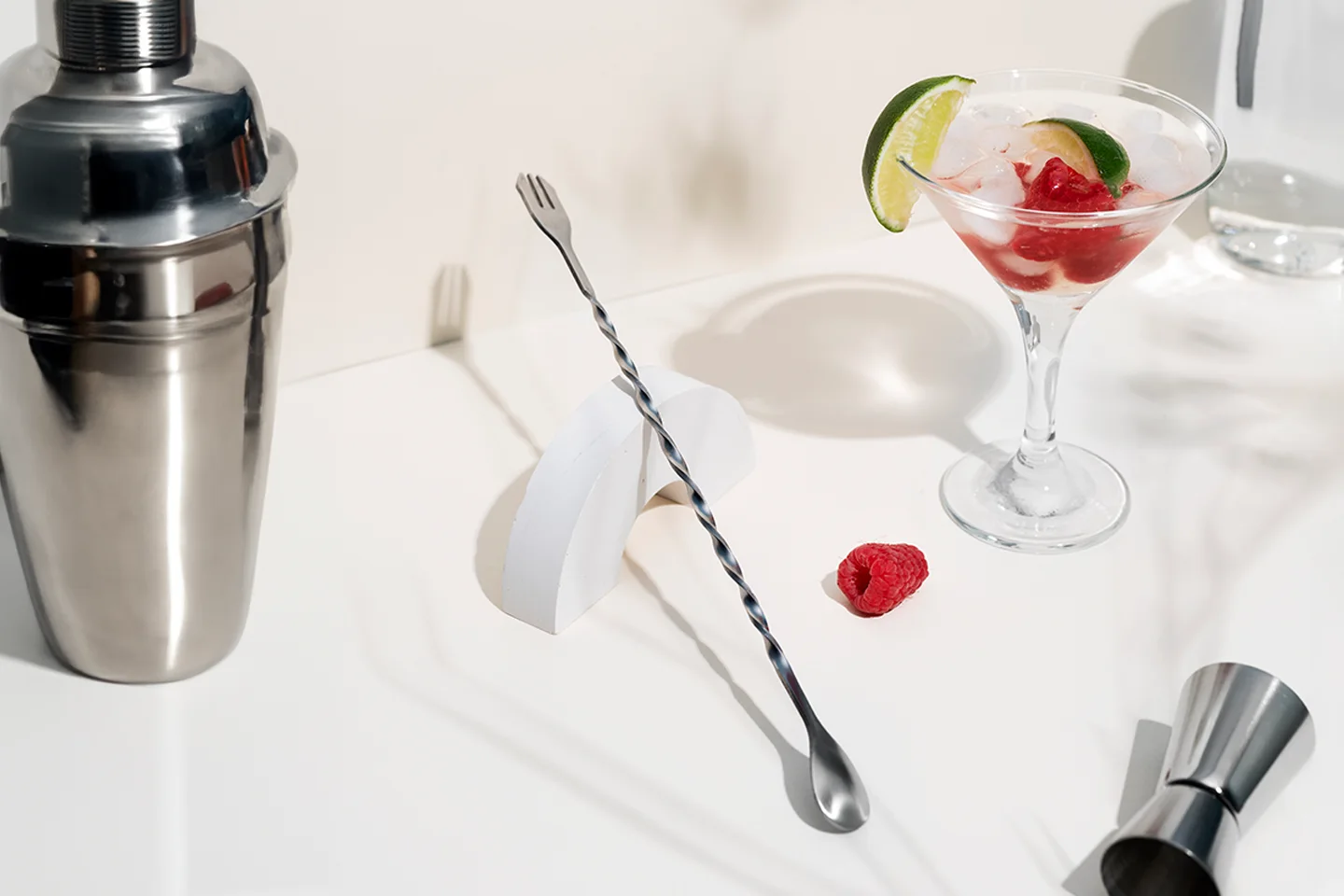 hi-Dry_grundaustattung Ein Cocktail-Shaker, ein Barlöffel, ein Jigger und ein Cocktailglas mit einem Getränk und frischen Himbeeren sowie Limettenscheiben auf einem weißen Hintergrund.