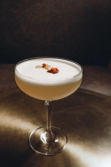 Ein Cocktail in einem Coupette-Glas. Die Flüssigkeit ist hellgelb und obenauf befindet sich eine weiße Schaumkrone. Als Garnitur sind rote Früchte zu sehen.