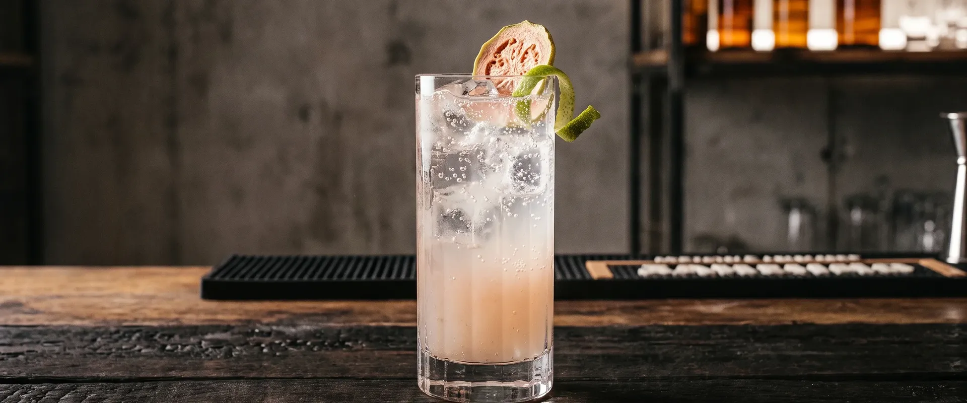 Platzhalter_1920x800 Rauchiger Guaven Fizz in einem hohen Glas mit Eiswürfeln, garniert mit Guavenscheibe und Limettenzeste, auf einer dunklen Holzbar mit verschwommenen Spirituosenflaschen im Hintergrund.