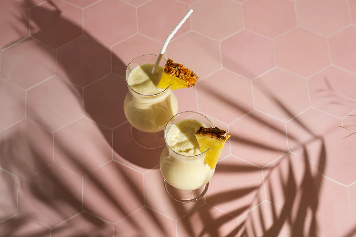 Zwei hohe Gläser mit einem cremig-gelben Cocktail, verziert mit frischen Ananasstücken. Die Gläser stehen auf einem pinken Tisch mit einem Schattenmuster von Palmblättern.