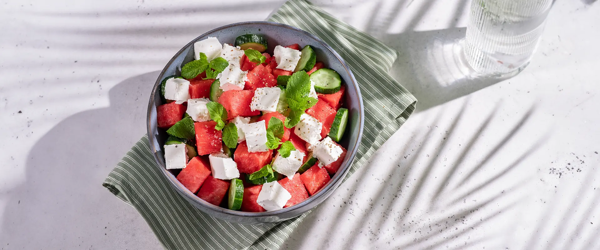Platzhalter_1920x800 Eine Schüssel mit einem bunten Salat aus gewürfelter Wassermelone, Gurkenscheiben, zerbröckeltem Feta und frischen Minzblättern, angerichtet auf einem grünen Stoffservietten.