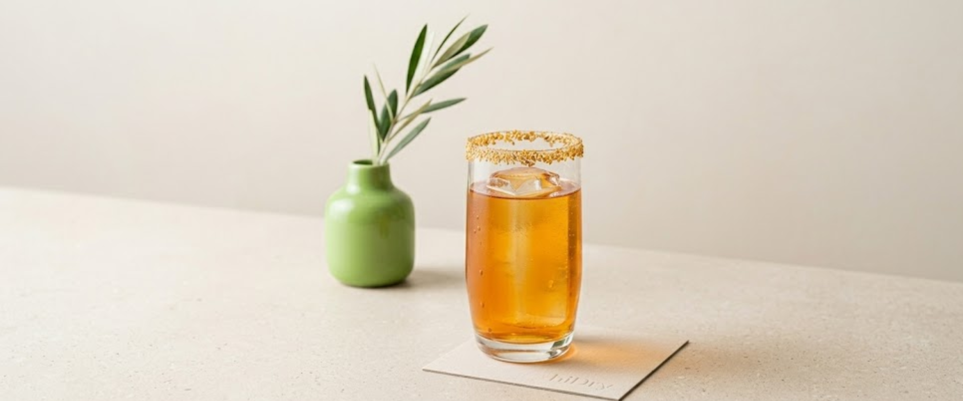 Platzhalter_1920x800 Goldener Open Sesam Eistee in einem hohen Glas mit gerösteten Sesamsamen am Rand und einem großen Eiswürfel, daneben eine grüne Vase mit Olivenzweig auf hellbeigem Hintergrund.